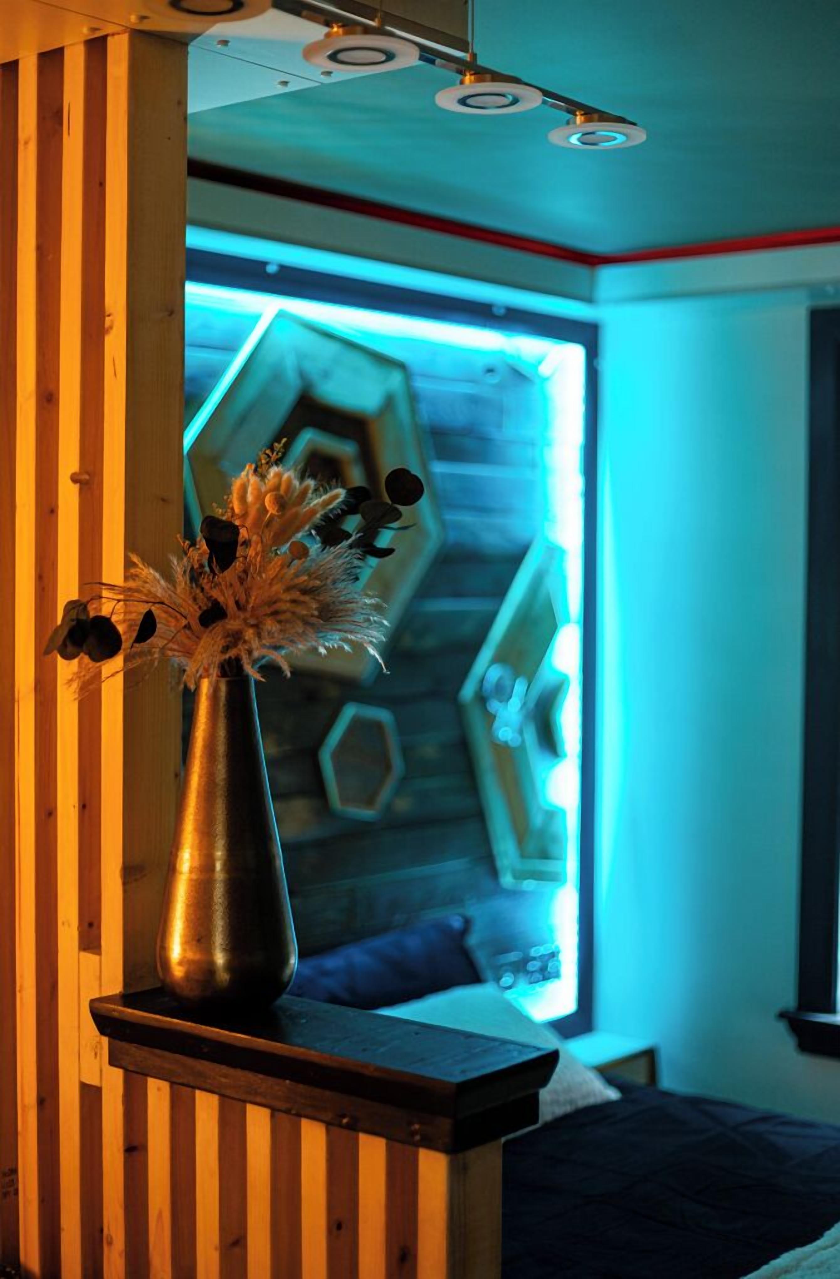 A room with a bed, a bedside table with a vase, and a window with a geometric pattern.