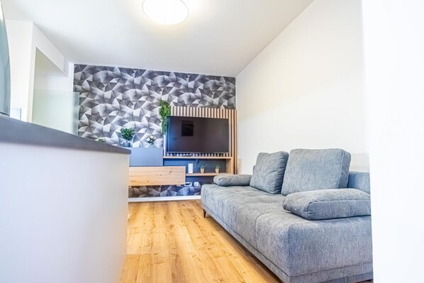Un salon moderne avec un canapé gris, un parquet en bois, un mur orné d’un motif géométrique et un meuble de rangement intégré pour les appareils multimédias.