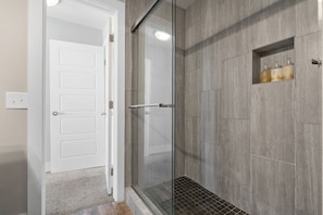 Et moderne badeværelse med en brusekabine i glas, vægge med grå fliser og en lille hylde med toiletartikler.