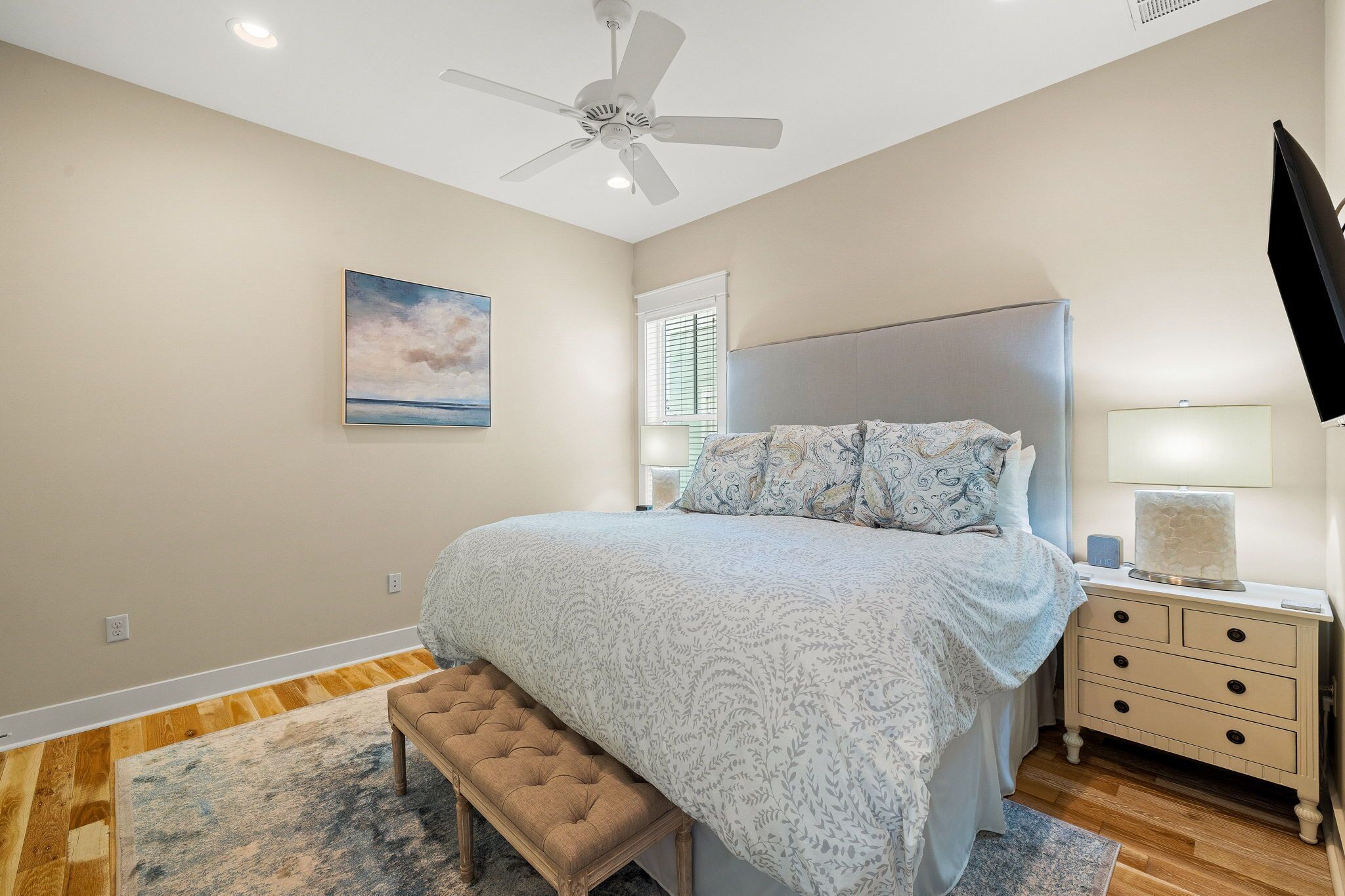 A bedroom with a bed, a ceiling fan, a nightstand, a bench, and a painting on the wall.