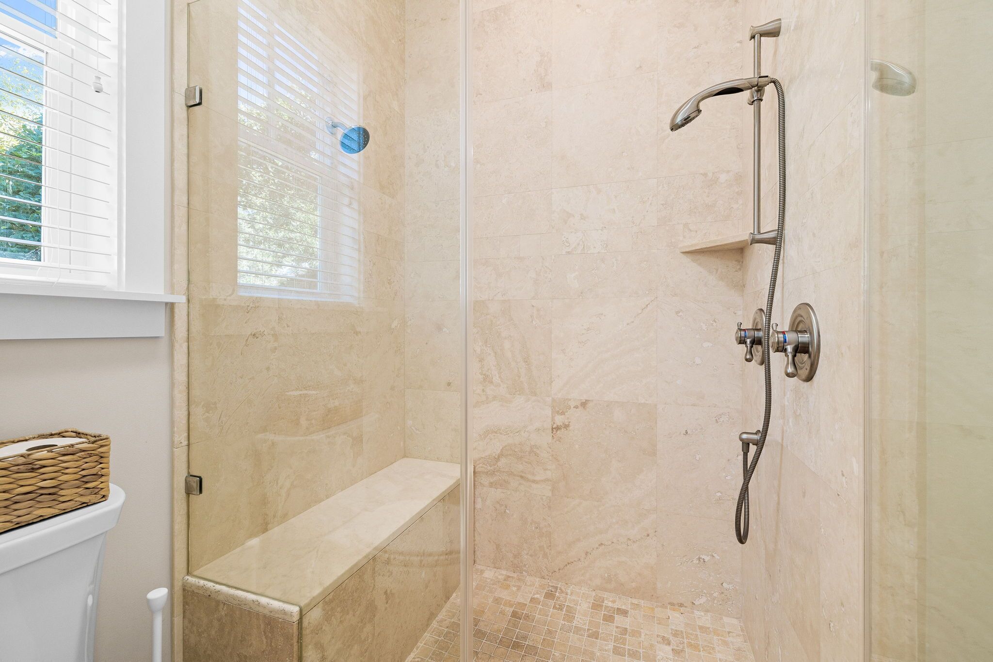 A shower with a handheld showerhead and a wall-mounted faucet, a bench, and a window with blinds.