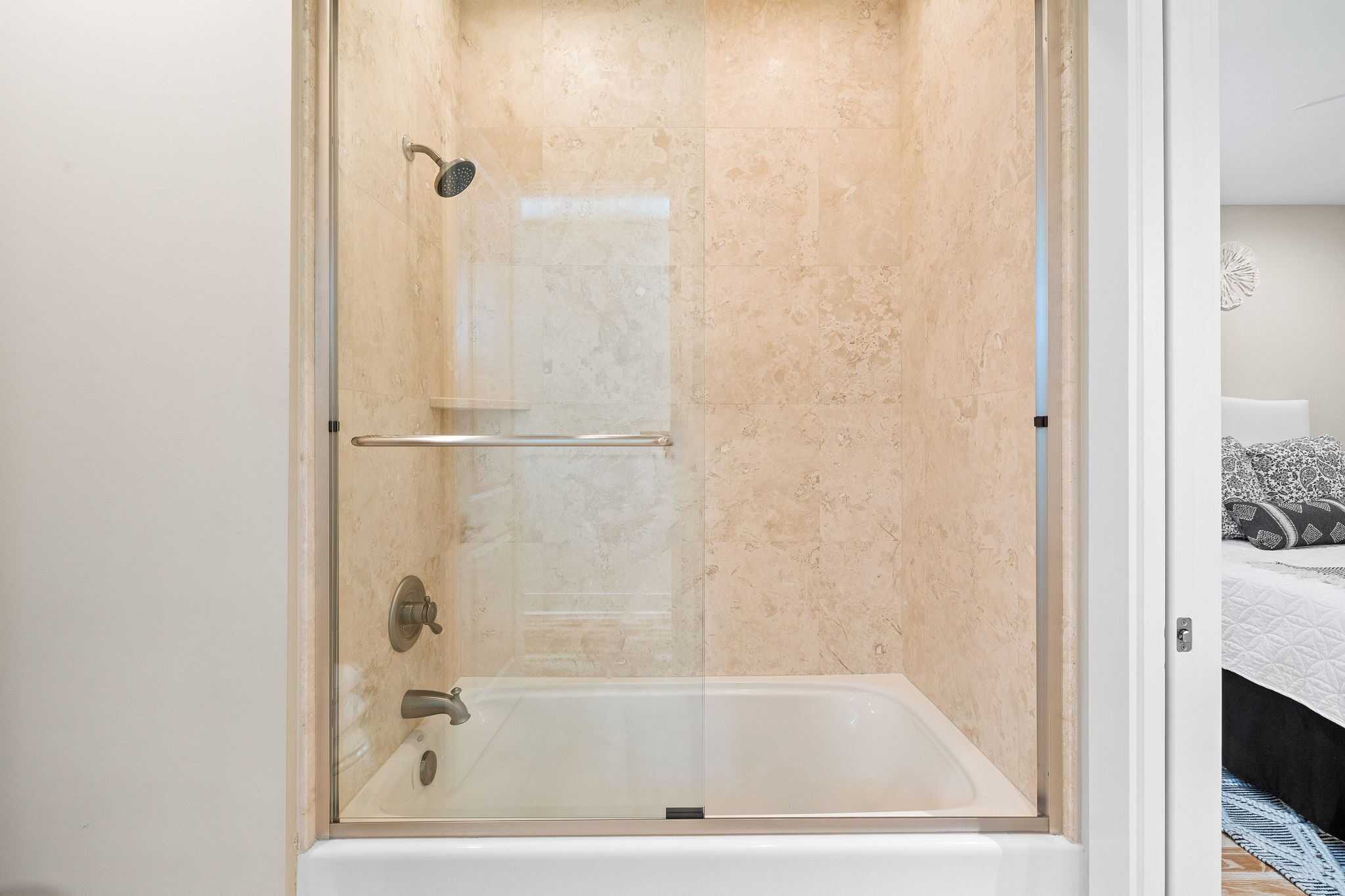 A glass-enclosed shower with a bathtub, beige marble walls, and a visible door leading to a bedroom with a bed and patterned pillows.