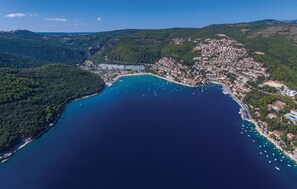 An aerial view of a coastal town with a marina, surrounded by hills and dense greenery.
