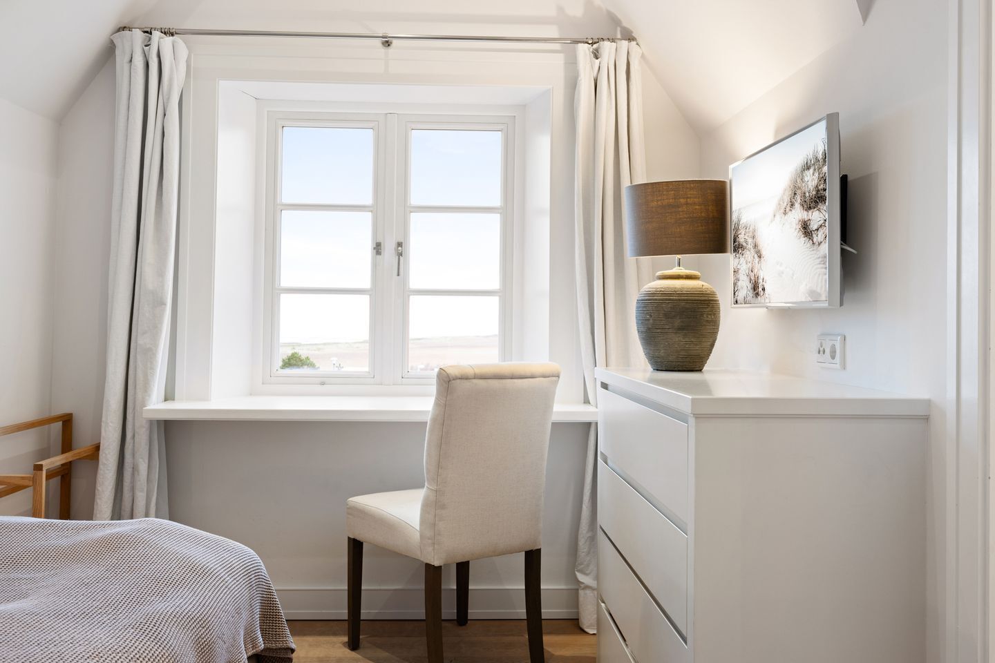 Un dormitorio con cama, una silla, un escritorio con lámpara y una ventana con cortinas.