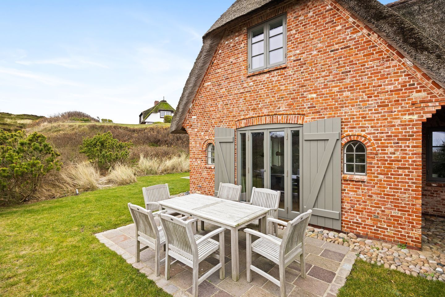 A brick house with a thatched roof, a patio with a table and chairs, and a view of the surrounding landscape.
