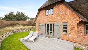Et murhus med stråtækt tag, træterrasse og en stenvæg.