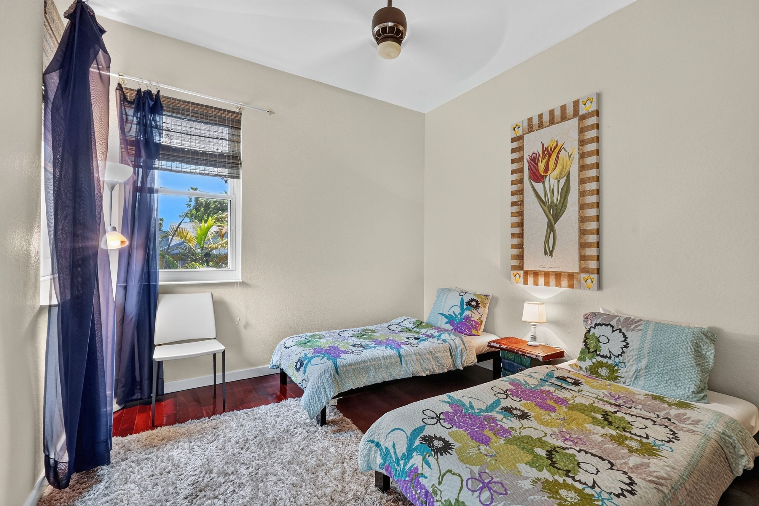 A bedroom with two beds, a window with a view, a ceiling fan, and a wall-mounted artwork.