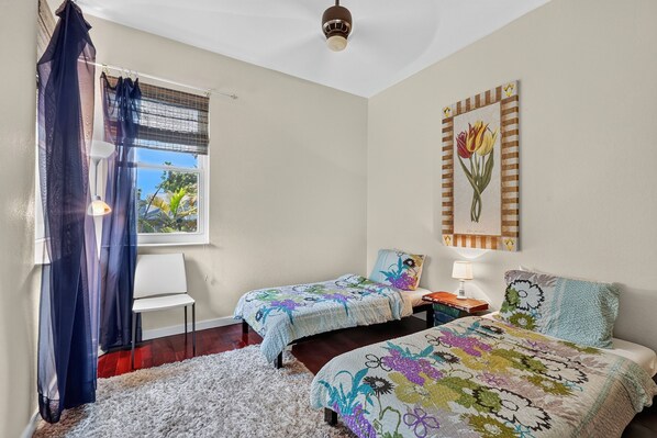 A bedroom with two beds, a window with a view, a ceiling fan, and a wall-mounted artwork.