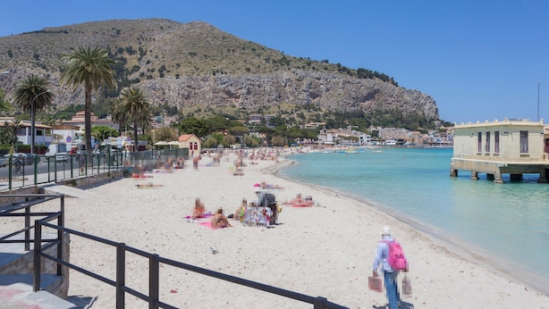 A beach with a rocky hillside in the background, a pier extending into the water, and a few people relaxing on the sand.