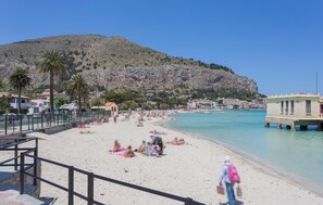 Uma praia com uma encosta rochosa ao fundo, um píer que se estende sobre a água e algumas pessoas relaxando na areia.
