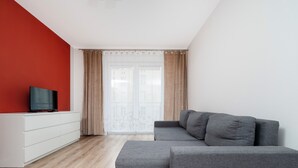 Un salon moderne avec un canapé d’angle gris, un tél éviseur à écran plat posé sur un meuble blanc et une grande fenêtre agrémentée de rideaux.
