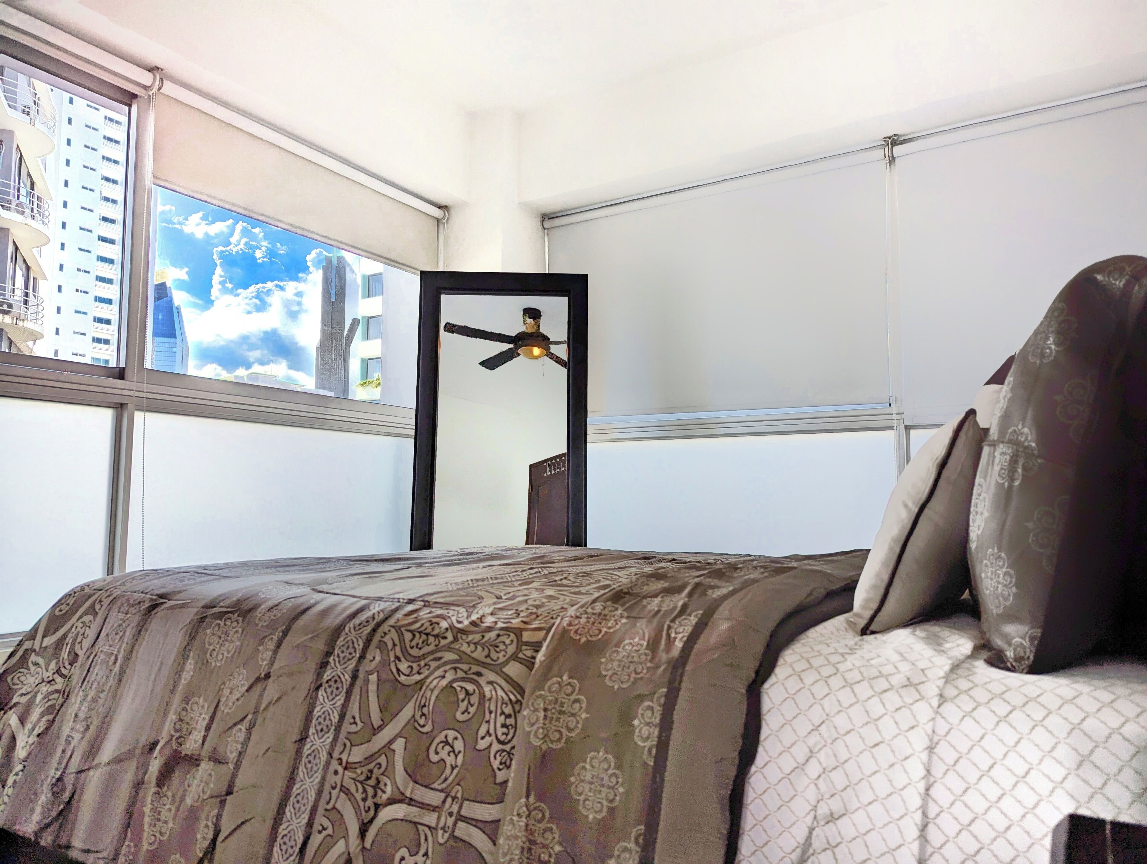A hotel room with a bed, a ceiling fan, a window with a view of buildings, and a door.