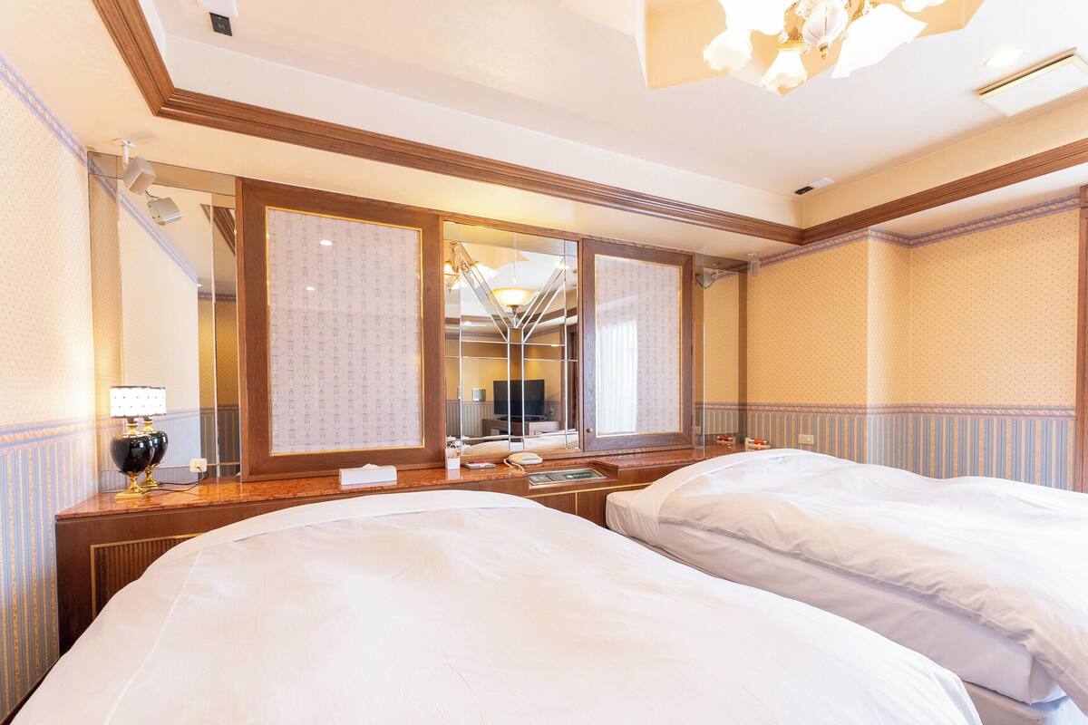 A hotel room with two beds, a large mirror, a desk with a lamp, and a television.