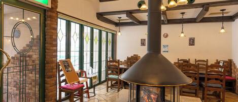 A restaurant interior with a stone-tiled floor, wooden chairs, a central wood-burning stove, and a green exit sign.