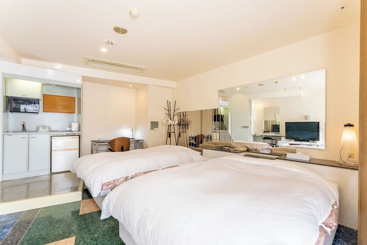 A hotel room with two beds, a desk with a chair, a TV, a mirror, and a kitchenette.