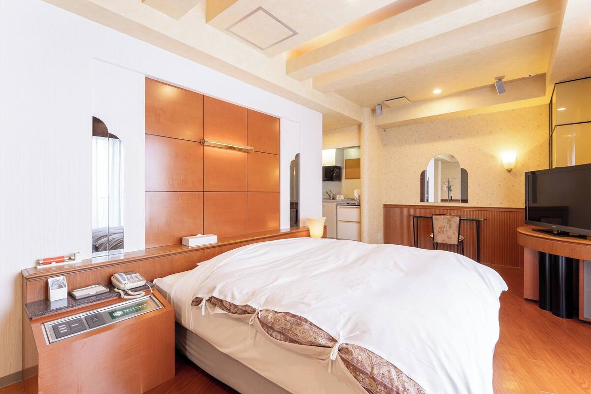 A hotel room with a large bed, a bedside table with a phone and a lamp, a TV, and a small kitchenette in the background.
