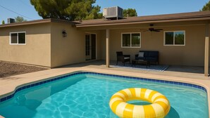 A swimming pool with a yellow inflatable ring, a covered patio with outdoor furniture, and a house with beige walls and a brown roof. - Desert Voyage | Pool | Near ASU + Old Town Scottsdale (Tempe)