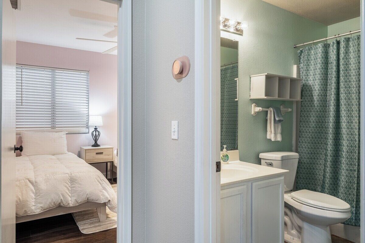 A bedroom with a bed, bedside table, and a window with blinds. Adjacent to the bedroom is a bathroom with a toilet, sink, and shower curtain.