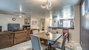 A modern living room with a wooden dining table, comfortable seating, a flat-screen TV, and a kitchen area with a bar.