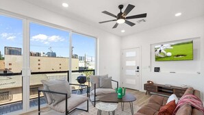 A modern living room with a large window, a ceiling fan, a flat-screen TV, and a sofa. - Home Run! Walk to Chase Field | 2BR Summer Stay in Downtown PHX (Phoenix)