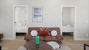 A modern living room with a brown leather sofa, two baseball-themed pillows, a green water bottle, and a marble-patterned coffee table. There are two doors leading to bedrooms with white bedding and blue-striped blinds. - Home Run! Walk to Chase Field | 2BR Summer Stay in Downtown PHX (Phoenix)
