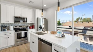 A modern kitchen with white cabinets, stainless steel appliances, and a large window offering a view of the city. - Home Run! Walk to Chase Field | 2BR Summer Stay in Downtown PHX (Phoenix)