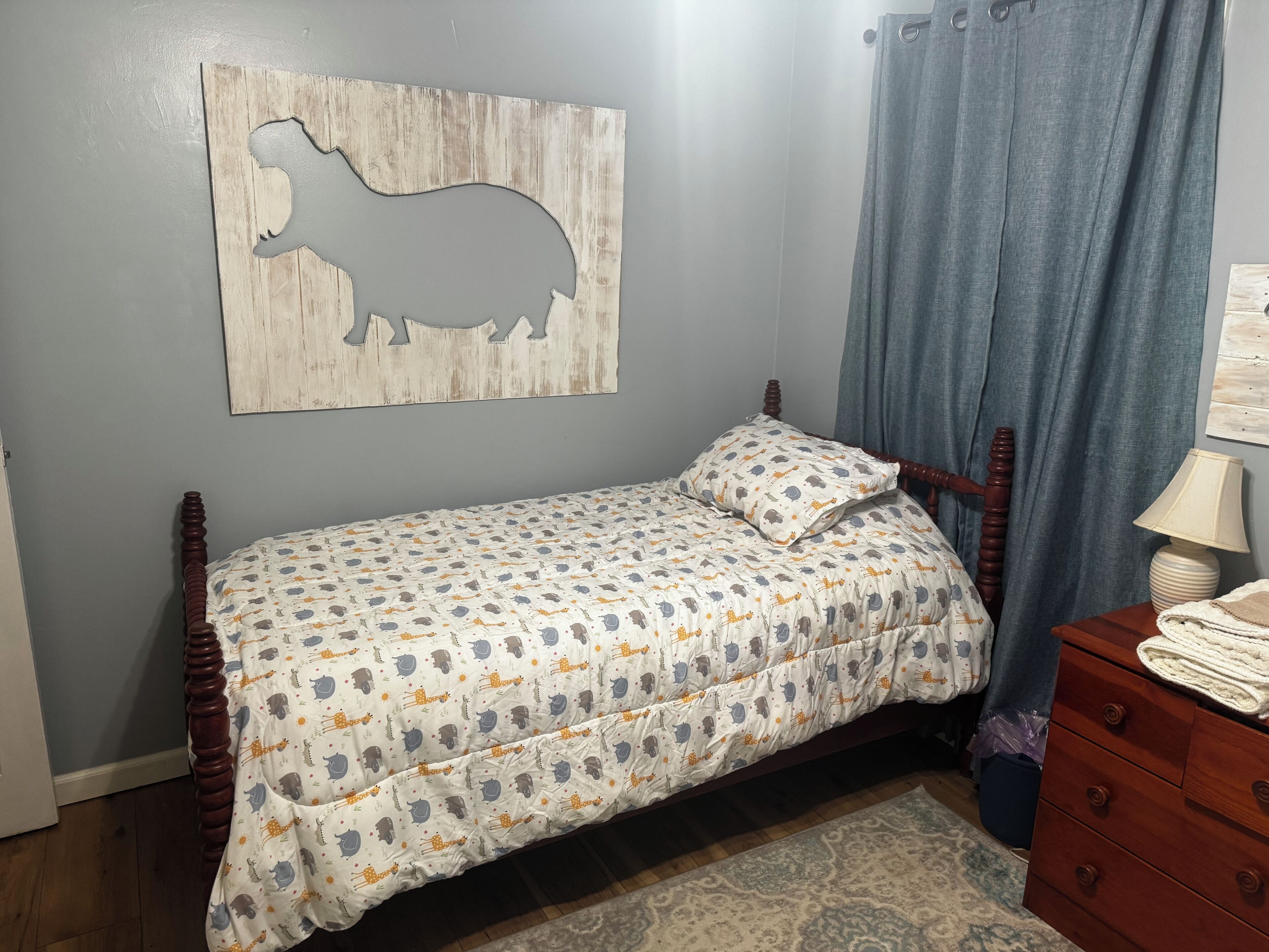 Een slaapkamer met een bed, een houten hoofdbord, een wanddecoratie van een varken, een blauw gordijn, een houten dressoir en een lamp.