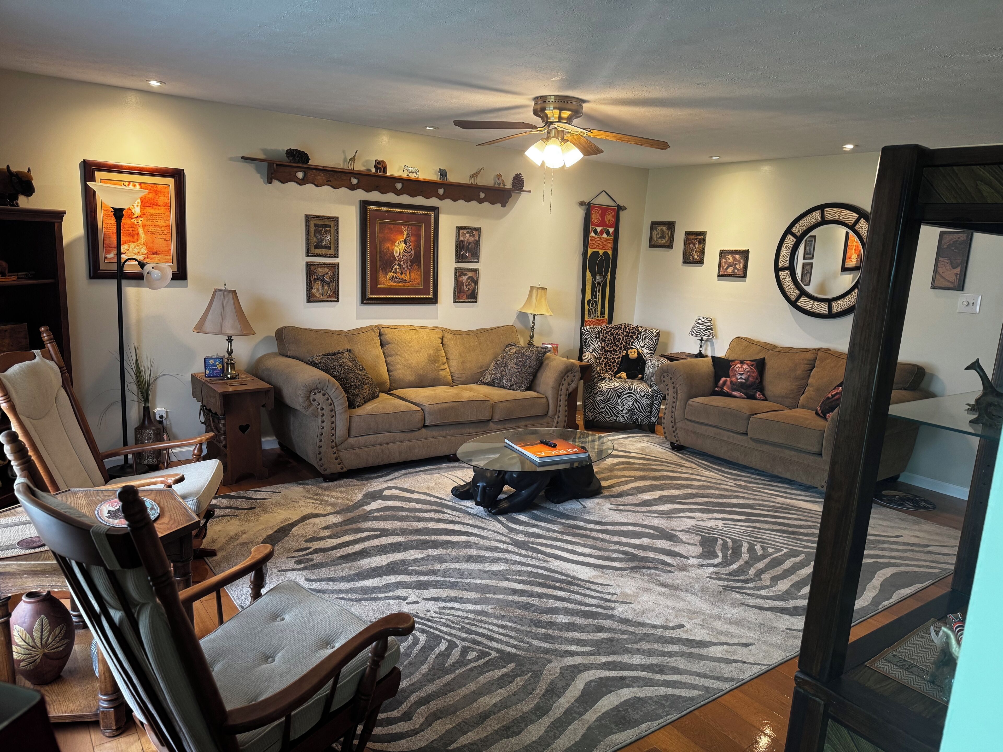 Un salon comprenant un canapé, des fauteuils, un tapis à motifs zébrés, un ventilateur de plafond et des décorations murales.