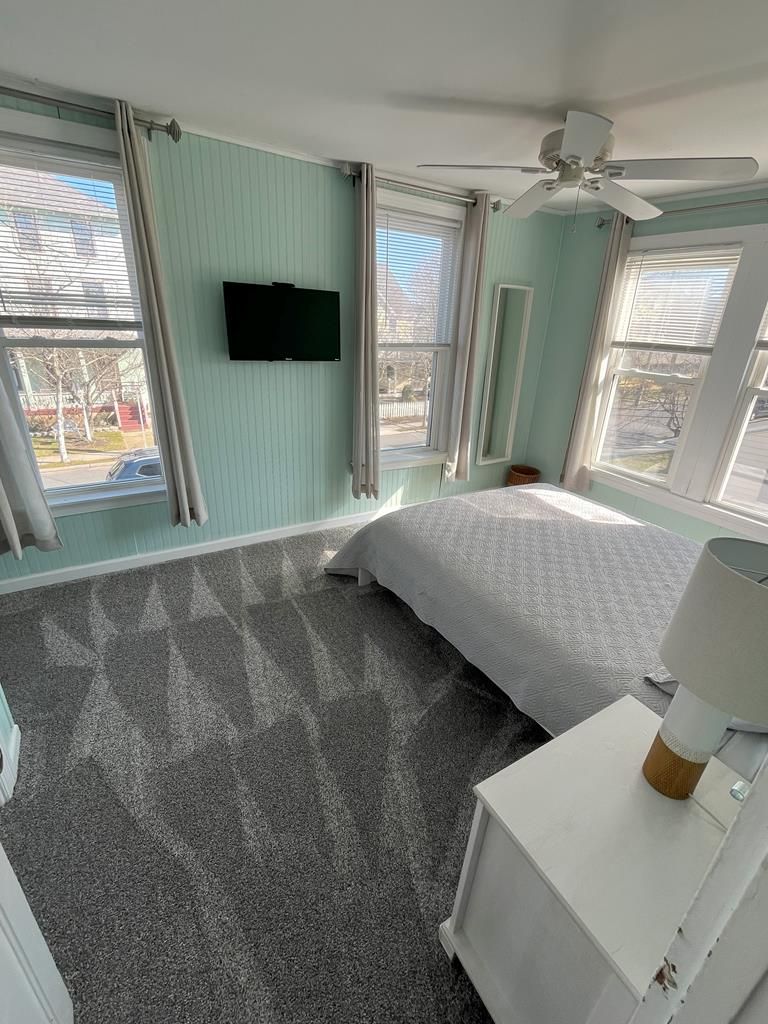 A bedroom with a bed, a ceiling fan, a flat-screen TV, and large windows.