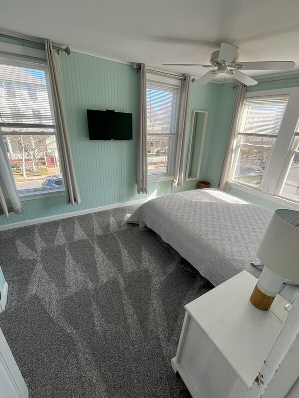 A bedroom with a bed, a ceiling fan, a flat-screen TV, and large windows.