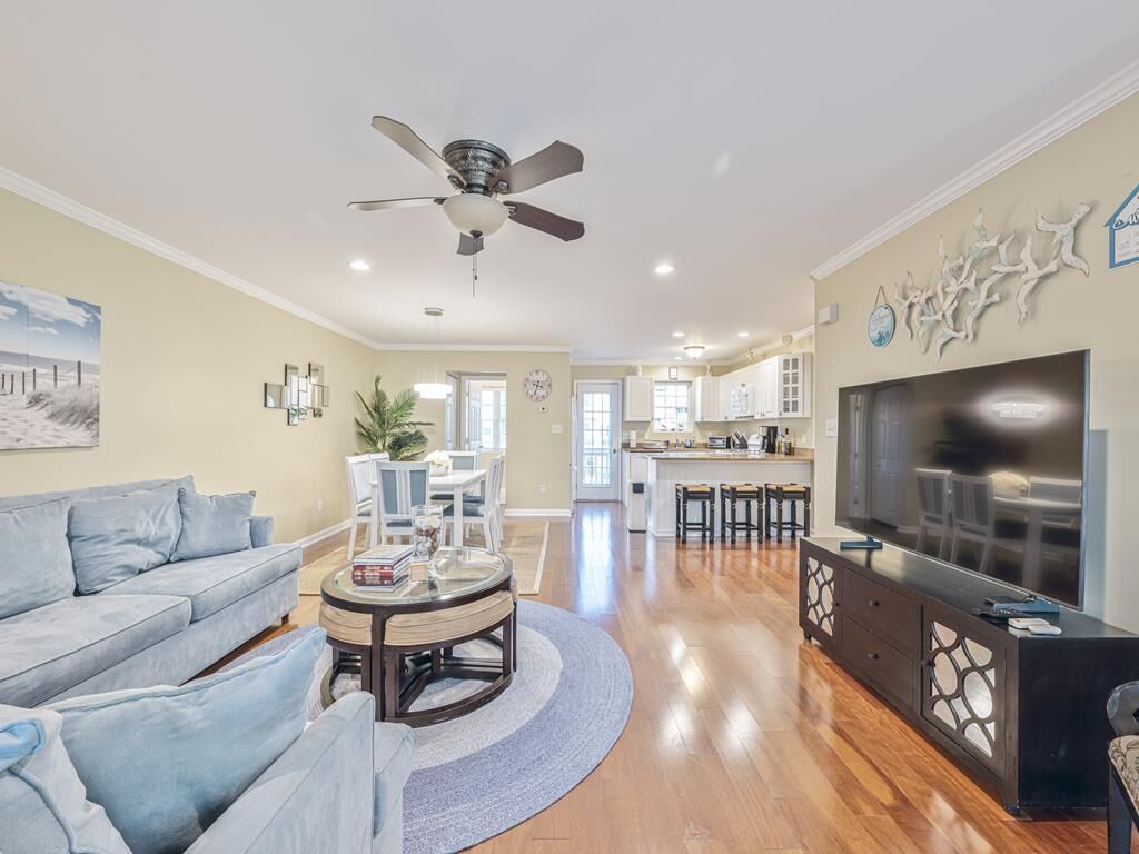 A spacious living room with a sofa, coffee table, and a TV. There is a ceiling fan, a kitchen area, and a dining area visible in the background.