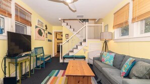 A living room with a TV, a wooden coffee table, a sofa with colorful cushions, a ceiling fan, and a staircase leading to another level.
