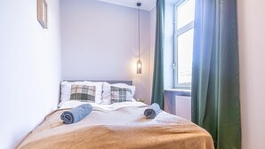 Una cama bien tendida con una manta marrón y dos toallas enrolladas, un aplique en la pared y una ventana con cortinas verdes.