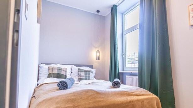 Una cama bien hecha con una manta y toallas enrolladas, una lámpara colgante y una ventana con cortinas.