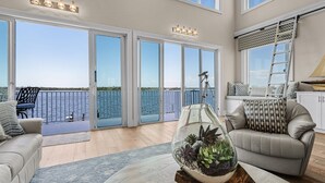 Ein geräumiges Wohnzimmer mit einem großen Glasfenster, das auf einen Balkon mit Meerblick führt, einer Glaskugel auf einem Holztisch und einem Ledersofa mit gemusterten Kissen.