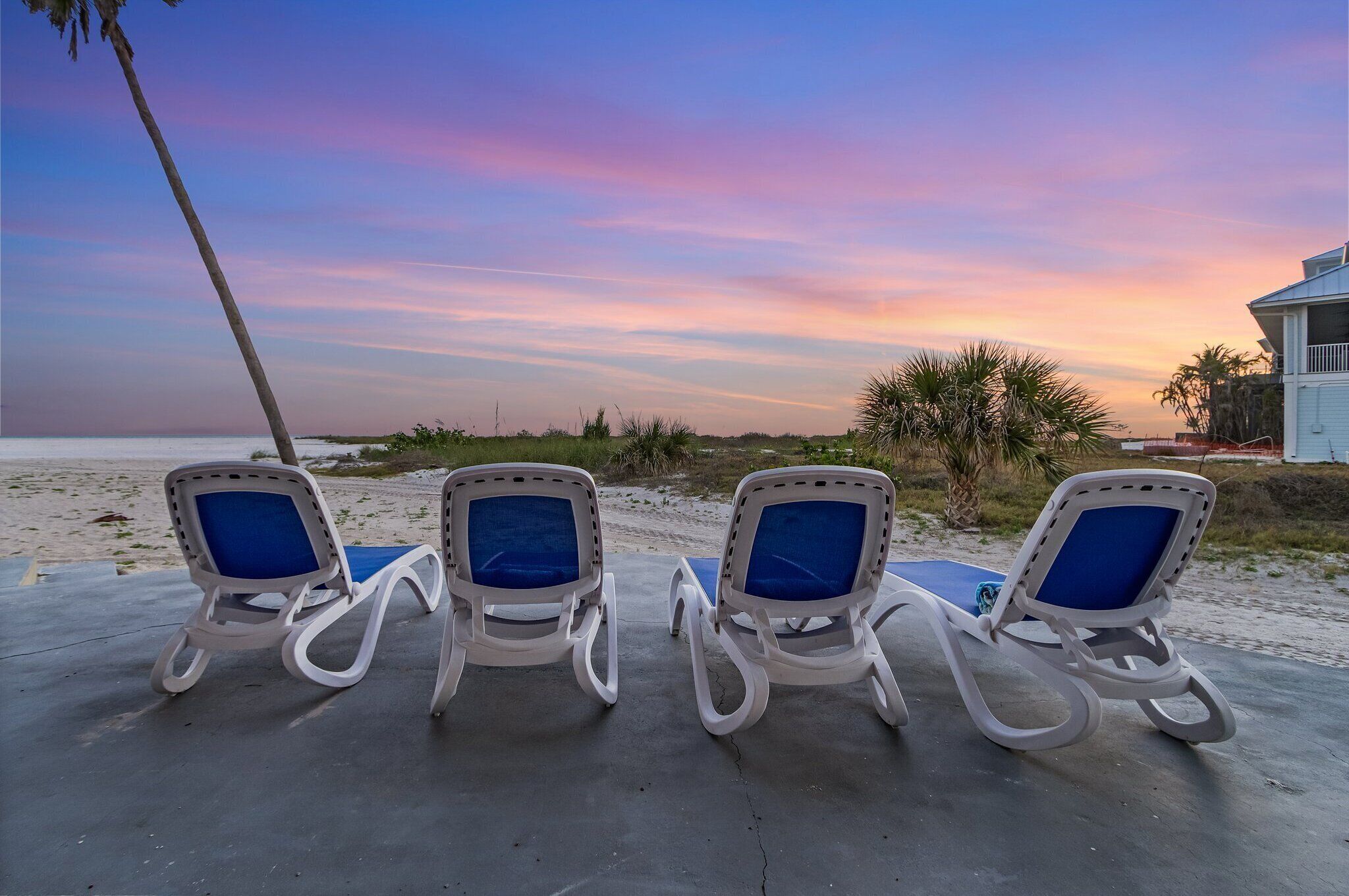 Four lounge chairs with blue cushions on a concrete surface, overlooking a beach with a sunset sky.