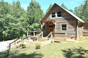 A log cabin with a porch, a small staircase, and a view of a forested area.