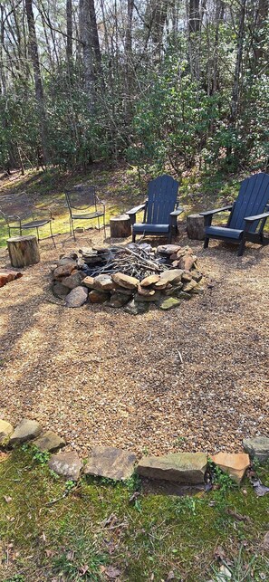 A fire pit surrounded by rocks and gravel, with Adirondack chairs and a metal fence in a wooded area. - Cozy Cabin w/Loft Bedroom * Firepit * Creek Access (Dahlonega)