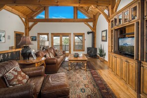 A spacious living room with a large wooden ceiling, leather furniture, a television, and a view of the outdoors through large windows.