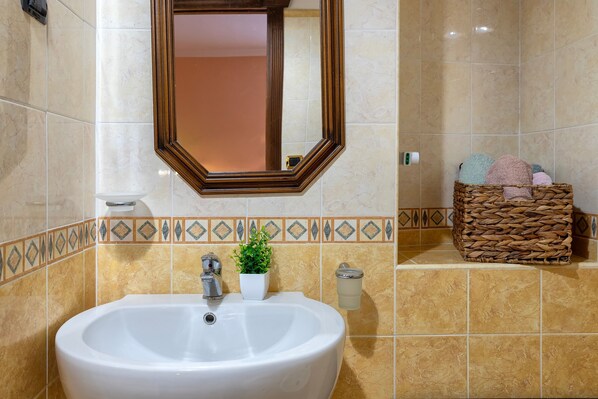A bathroom with a white sink, a mirror, a potted plant, a wicker basket with towels, and a soap dispenser.