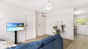 Ein modernes Wohnzimmer mit einem blauen Sofa, einem Flachbildfernseher, der eine Strandansicht zeigt, einem Deckenventilator und einem durch eine offene Tür einsehbaren Küchenbereich.