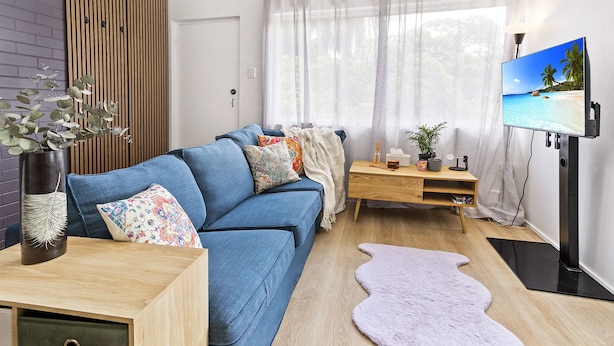 Ein modernes Wohnzimmer mit einem blauen Sofa, einem Holztisch, einem Fernseher auf einem Standfuß und Blick auf einen Strand durch transparente Vorhänge.