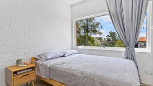 Ein Schlafzimmer mit weißer Ziegelwand, einem hölzernen Bettgestell, einem Nachttisch mit Schubladen, einem Fenster mit Vorhängen und Blick auf Bäume und Gebäude.