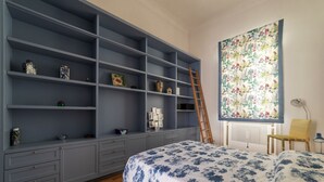 A bedroom with a large grey wardrobe, a bed with a floral bedspread, a ladder leaning against the wardrobe, and a floral tapestry on the wall.