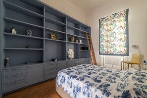 A bedroom with a large grey wardrobe, a bed with a floral bedspread, a ladder leaning against the wardrobe, and a floral tapestry on the wall.