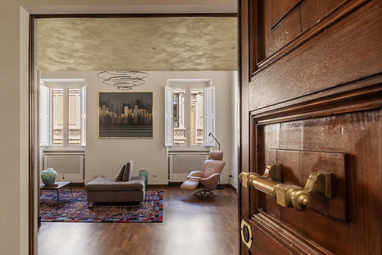 Ein Wohnzimmer mit einer Couch, einem Sessel und einem farbenfrohen Teppich. An der Wand hängt ein Gemälde und ein Fenster ist mit Jalousien versehen.