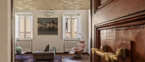 A living room with a sofa, armchair, and a colorful rug. There is a painting on the wall and a window with blinds.