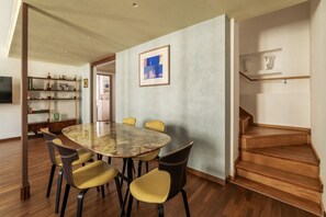 A dining area with a marble table and yellow chairs, a bookshelf, a framed artwork, and a wooden staircase.