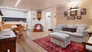 Ein gemütlicher Wohnbereich mit einer Couch, Hocker und einem roten Teppich. Außerdem sind ein Holzschrank und ein Sessel zu sehen.