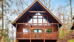 A two-story log cabin with a balcony, a green door, and a clear view of the surrounding forest.